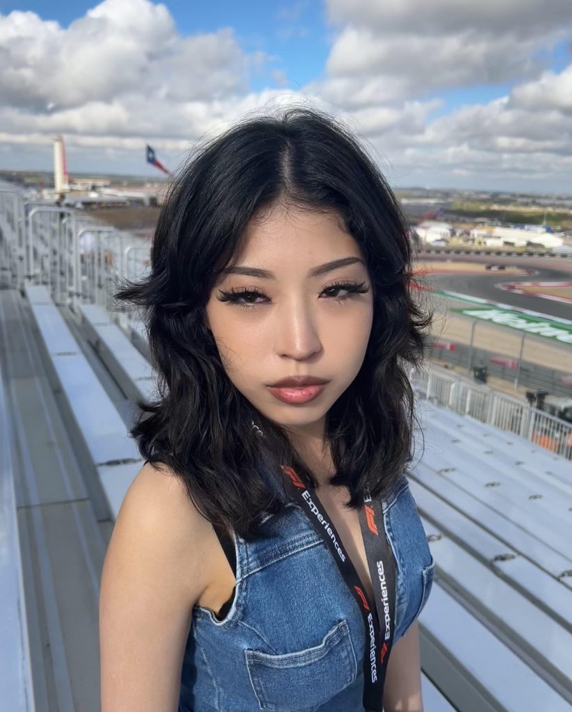 Meet The Team: A woman with dark hair and a denim top stands in front of empty stadium seats at a racetrack on a partly cloudy day.