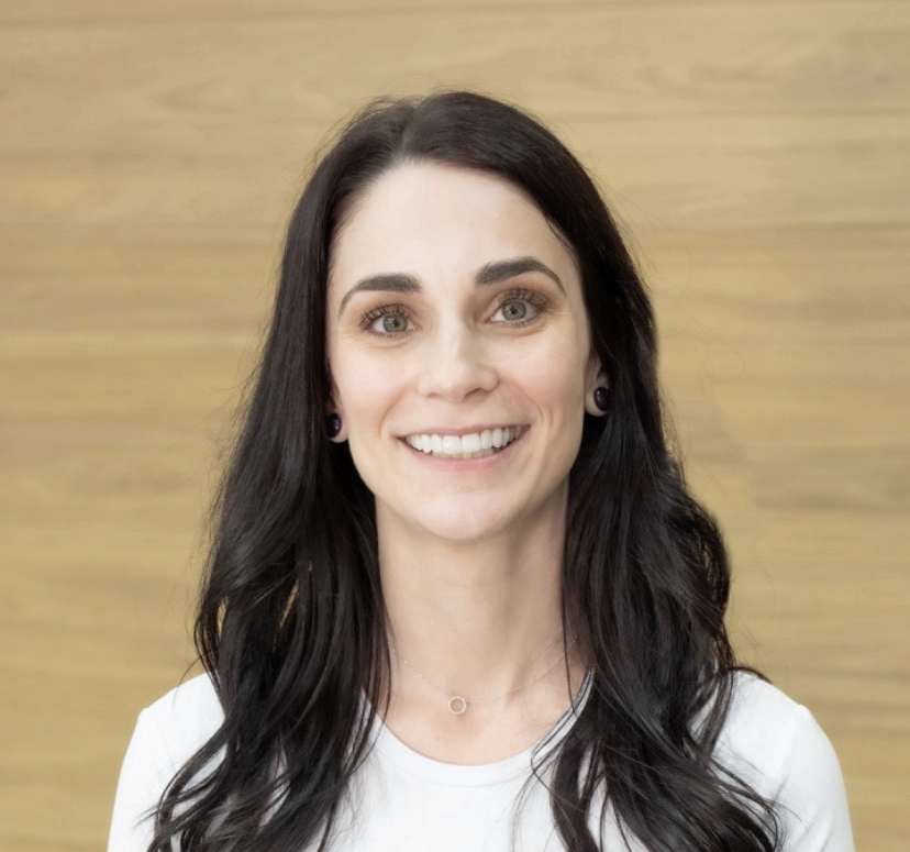 Meet the team: A woman with long dark hair and fair skin smiles at the camera, standing in front of a light wood background. She is wearing a white top and a small necklace.