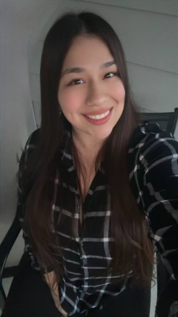 Meet the Team: A woman with long dark hair, wearing a black and white plaid shirt, sits on a chair and smiles at the camera.