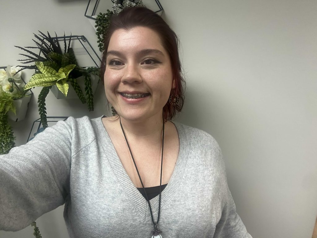 Meet The Team: A woman with reddish-brown hair wearing a light gray sweater smiles at the camera. Green plants in geometric wall planters are visible in the background.
